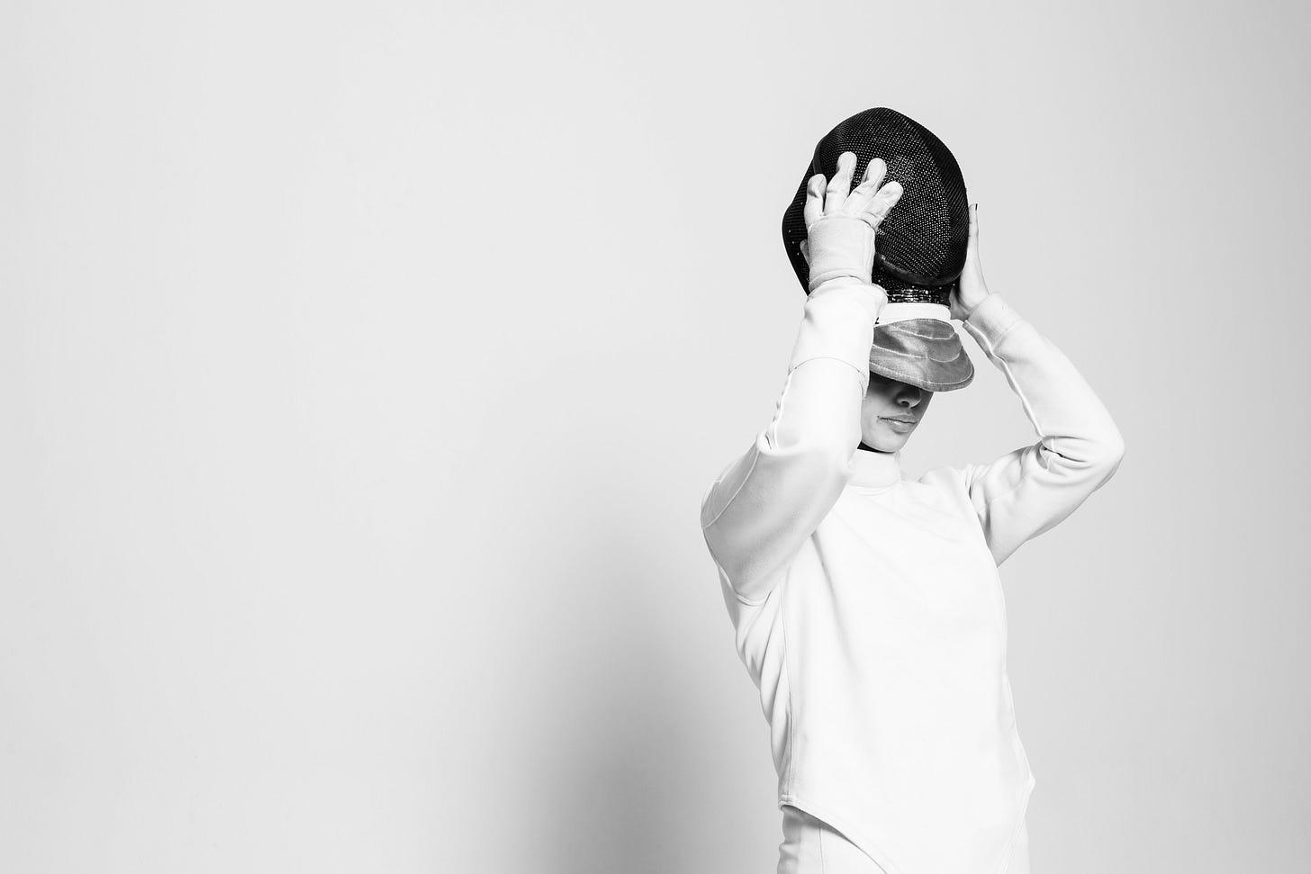 A black and white image. To the right is a fencer, in all white, taking off her fencing mask.