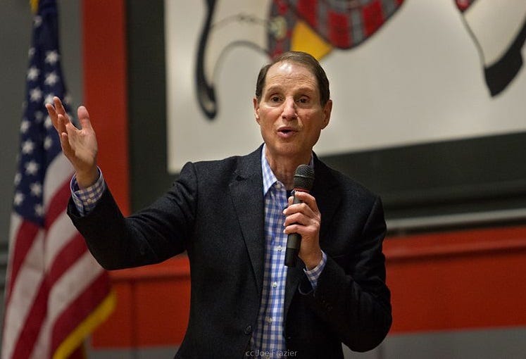 Ron Wyden, wearing a jacket, blue and white checked shirt, and no tie, gestures as he speaks into a microphone at a high school gymnasium