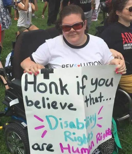 Emily sitting in a power wheelchair, holding a hand painted sign that reads "Honk if you believe that disability rights are human rights." Emily sitting in a power wheelchair, holding a hand painted sign that reads "Honk if you believe that disability rights are human rights."