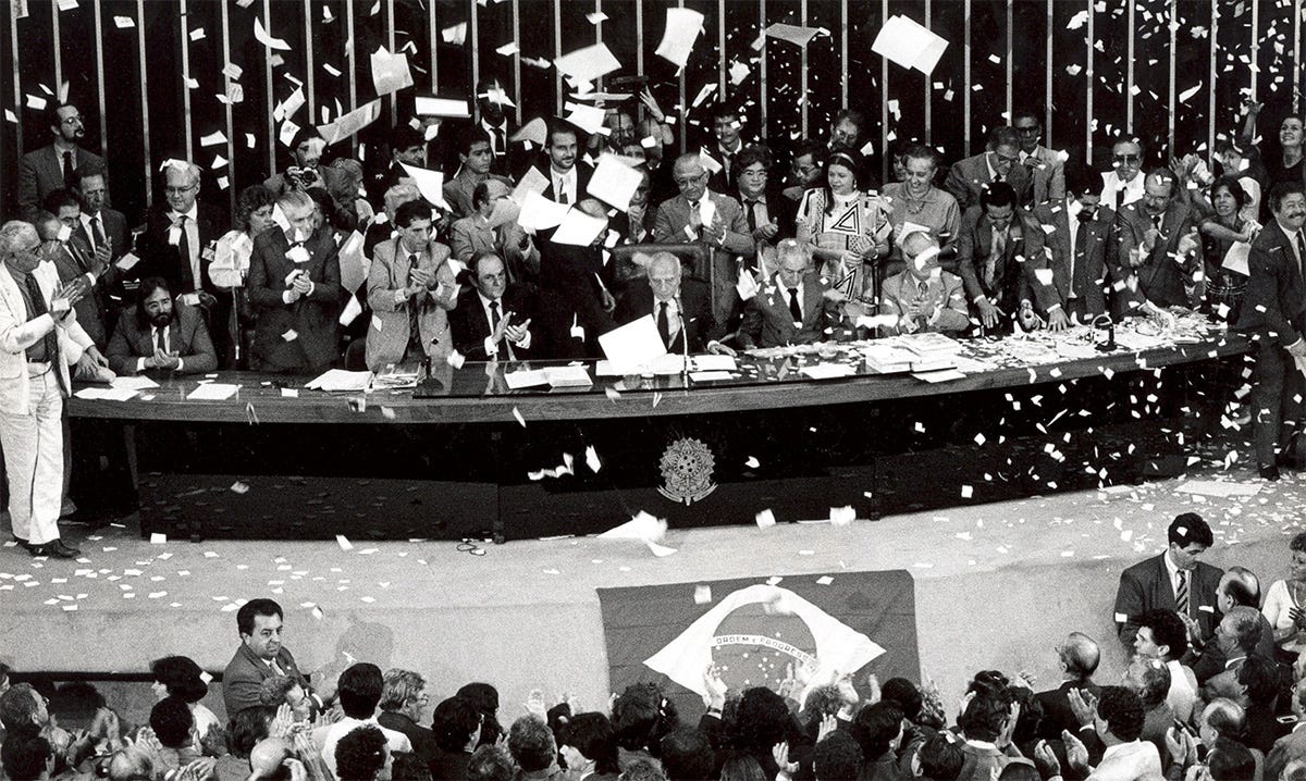 Fotografia histórica em preto e branco capturada de um ângulo alto, registrando o Plenário da Câmara dos Deputados em Brasília totalmente tomado por uma celebração festiva durante a promulgação da Constituição de 1988. A imagem é marcada por uma intensa "chuva" de centenas de pedaços de papéis brancos recortados e folhas de documentos que flutuam pelo ar, cobrindo o teto, a bancada e o chão como confetes. No centro, atrás de uma longa mesa diretora curva e escura adornada com o brasão da República, dezenas de parlamentares, políticos e autoridades vestindo paletó e gravata aplaudem e comemoram de pé. No quadrante inferior, uma numerosa plateia acompanha o evento de costas para a câmera, também aplaudindo em direção à mesa principal. À frente dessa plateia inferior, estendida no chão da rampa que dá acesso ao palanque, destaca-se uma bandeira do Brasil de tamanho médio. A composição transmite uma atmosfera de grande comoção coletiva, euforia política e saturação visual provocada pelos papéis em movimento no ar. Fotografia histórica em preto e branco capturada de um ângulo alto, registrando o Plenário da Câmara dos Deputados em Brasília totalmente tomado por uma celebração festiva durante a promulgação da Constituição de 1988. A imagem é marcada por uma intensa "chuva" de centenas de pedaços de papéis brancos recortados e folhas de documentos que flutuam pelo ar, cobrindo o teto, a bancada e o chão como confetes. No centro, atrás de uma longa mesa diretora curva e escura adornada com o brasão da República, dezenas de parlamentares, políticos e autoridades vestindo paletó e gravata aplaudem e comemoram de pé. No quadrante inferior, uma numerosa plateia acompanha o evento de costas para a câmera, também aplaudindo em direção à mesa principal. À frente dessa plateia inferior, estendida no chão da rampa que dá acesso ao palanque, destaca-se uma bandeira do Brasil de tamanho médio. A composição transmite uma atmosfera de grande comoção coletiva, euforia política e saturação visual provocada pelos papéis em movimento no ar.