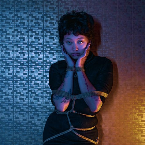 A girl dressed in black is posing with confidence, tied in ropes, over a blue and orange tiled background.