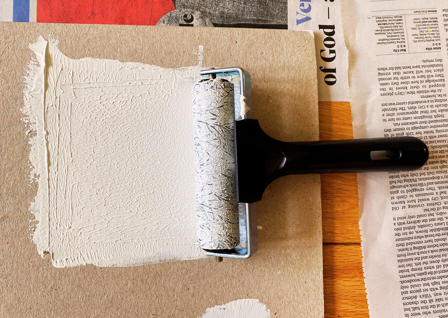photo from the printing process: a rubber roller, covered in white paint, rests on a sheet of cardboard, which is also covered in white paint. the cardboard rests on bits of overlapping newspaper – floor protection. The roller has just been used to ink the woodblock, so there are filigree patterns of the rubber below showing through the wet paint. 