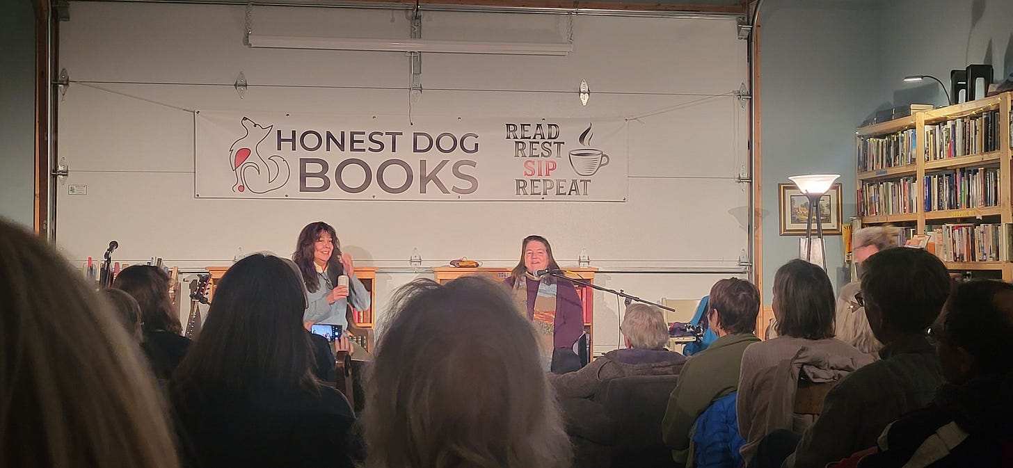 Deborah Taffa and Pam Houston sitting in chairs in front of a garage door with an Honest Dog Books sign on it. There are book shelves one side wall and people sitting in chairs in the audience facing the authors.