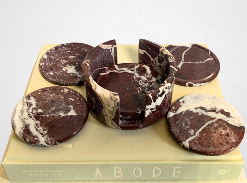 May include: A set of marble coasters and a holder. The coasters and holder are a deep burgundy color with white veining. The holder is a circular shape with a curved design. The coasters are round and sit around the holder.