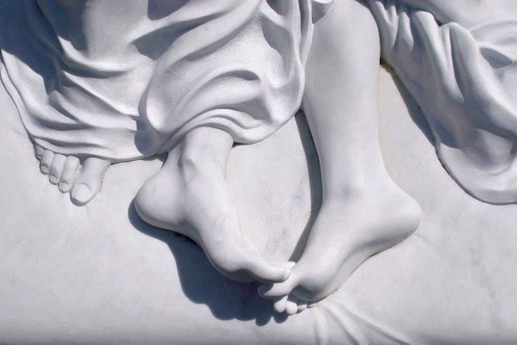 Close-up of a white marble sculpture showing two bare feet gently touching at the toes, with softly carved draped fabric surrounding the ankles.