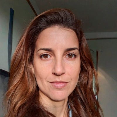 Retrato em primeiro plano de uma mulher de cabelos longos e castanhos, olhando para a câmera e sorrindo levemente, em um ambiente interno.