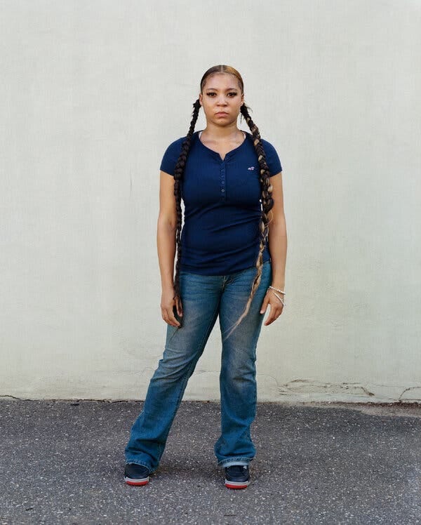 Najya Hannah-Jones, a teenage Black girl in a blue short-sleeved shirt and jeans, stands outdoors in front of a beige-painted wall. Her expression is serious. Her dark hair, some of it bleached blond, is pulled into two waist-long braids.