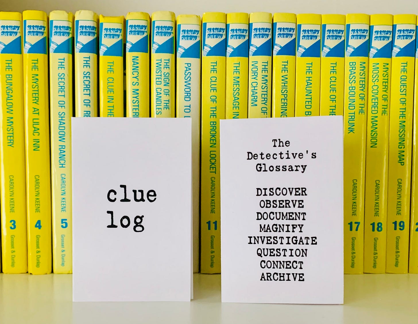 A zine called Clue Log rests on a shelf before a row of yellow Nancy Drew books