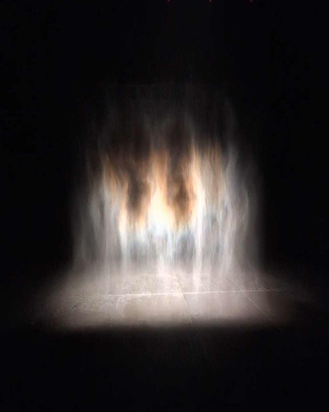 Abstract image of illuminated mist or water vapor in darkened space creating ghostly vertical streaks of white and warm amber light against black background, with visible floor line at bottom