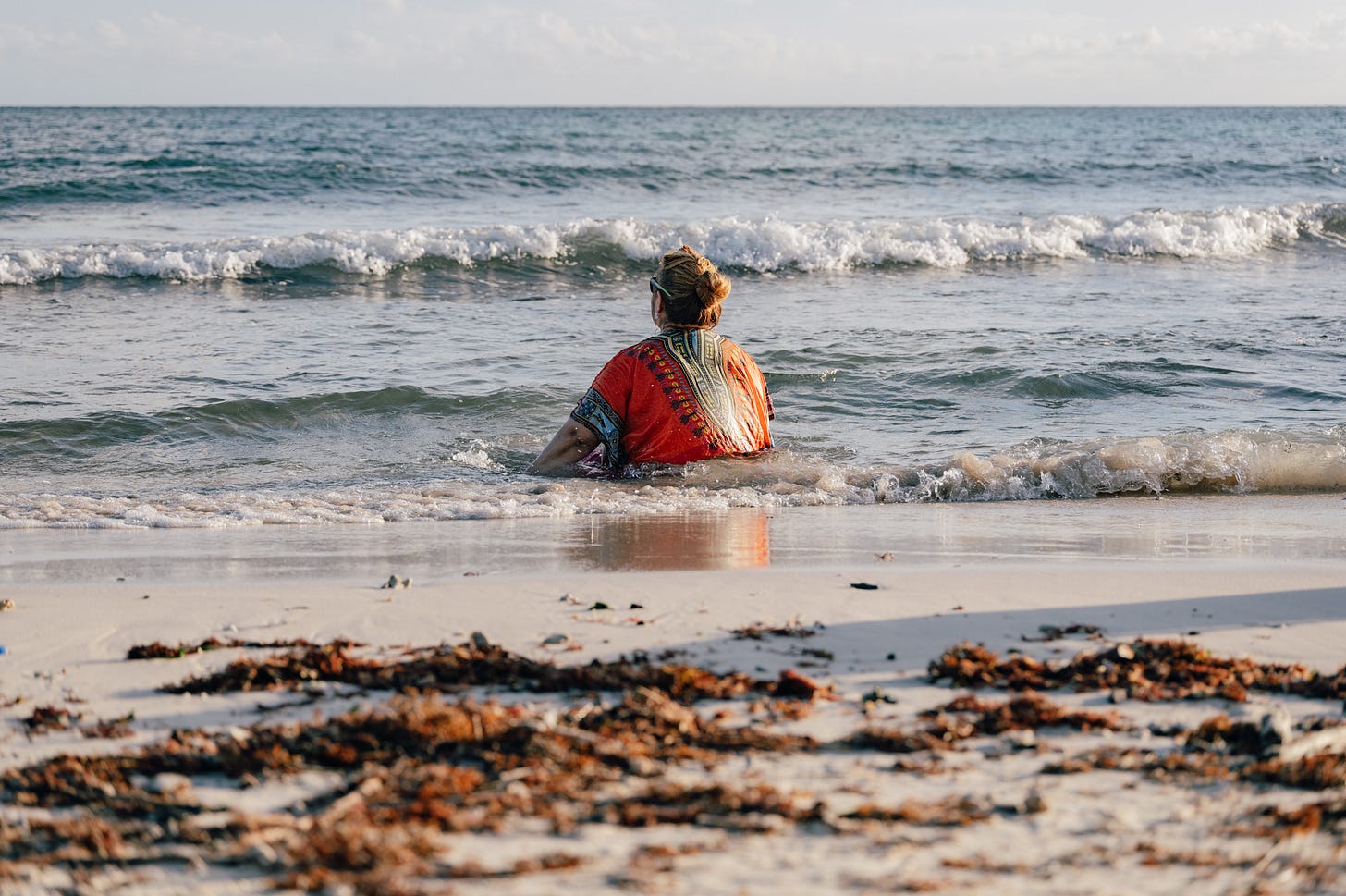 A woman in an orange kaftan sits in the shallow surf, her back to the camera, facing the open ocean. Seaweed is scattered across the sand in the foreground. A woman in an orange kaftan sits in the shallow surf, her back to the camera, facing the open ocean. Seaweed is scattered across the sand in the foreground.