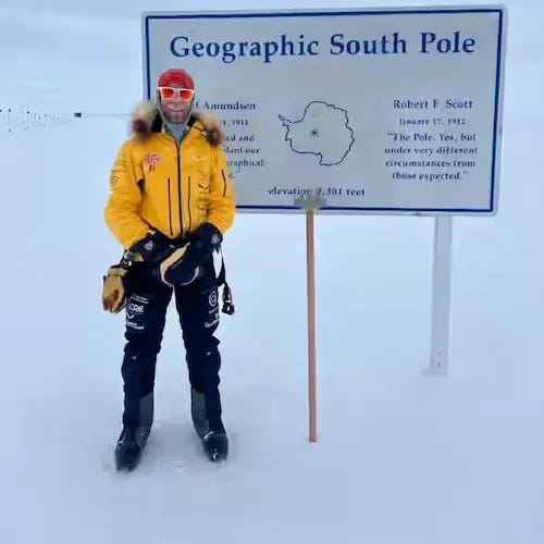 The image shows Jonny Huntington standing at the Geographic South Pole in Antarctica. He is dressed in heavy polar gear, including a bright yellow parka with fur trim on the hood, black pants, and gloves, clearly prepared for extreme cold weather conditions. He is also wearing protective goggles with a red tint. Behind them, there is a sign that reads "Geographic South Pole" and includes information about the location, such as the names Roald Amundsen and Robert F. Scott, as well as references to their historic expeditions. The area is covered in snow, emphasizing the remote and icy environment.
