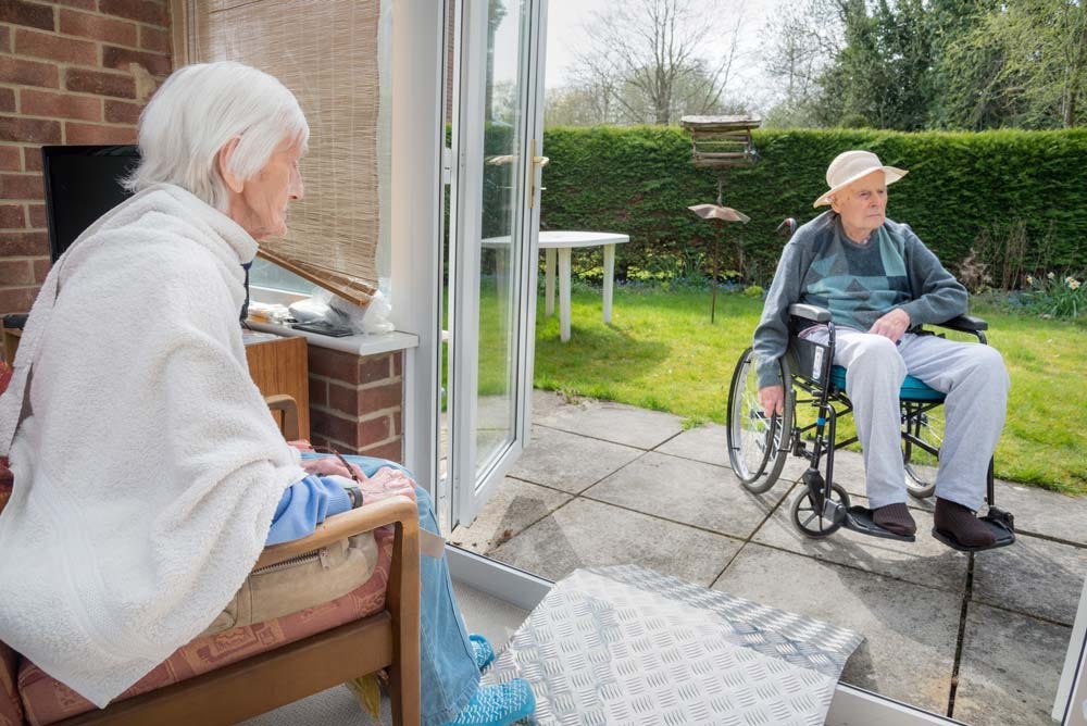 Image of an elder couple sitting on an outdoor patio enjoying the garden view; she sits in a chair while he sits in a wheelchair.