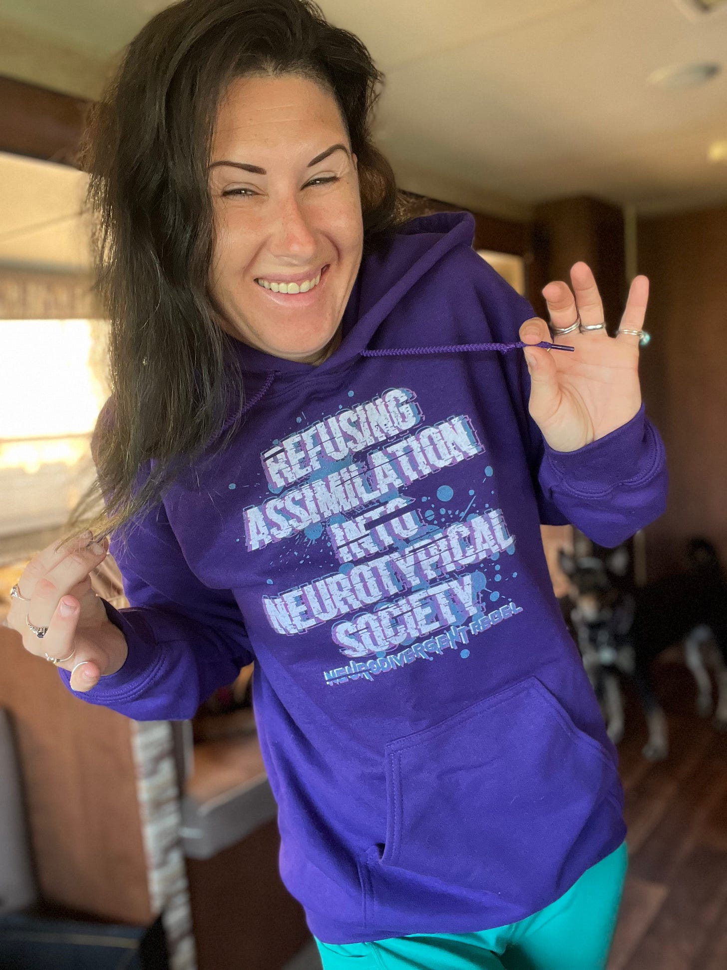 Lyric is wearing a purple hoodie and teal pants. On the hoodie is text in an apocalyptic font that reads: Refusing Assimilation into NeuroTypical Society in white and purple text, they are smiling for the photo and Bear, the dog, can be seen in the blurred background. 