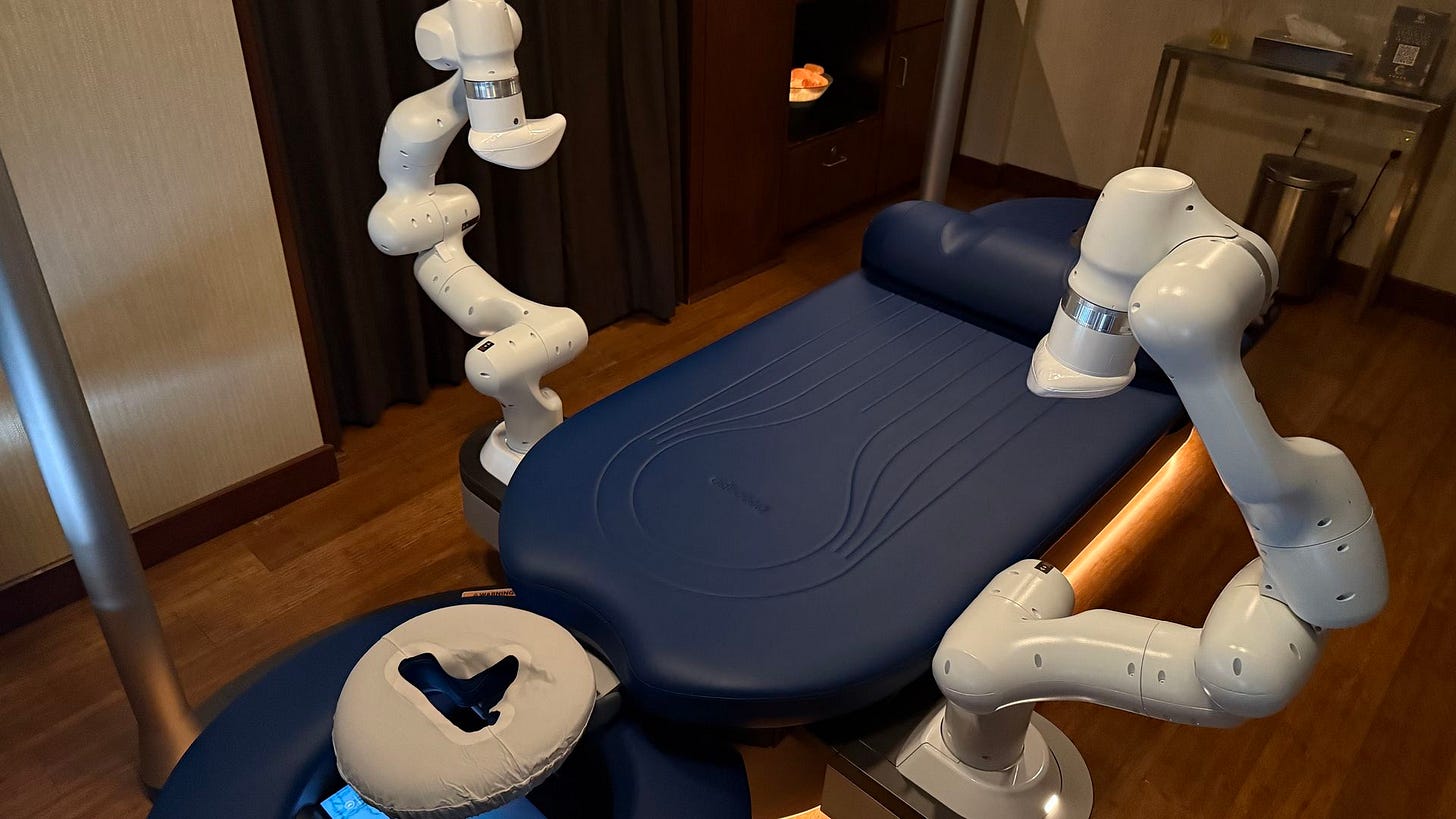 A futuristic massage or medical chair with blue cushions and two white robotic arms in a dimly lit room with wooden floor and a sink area in the background.