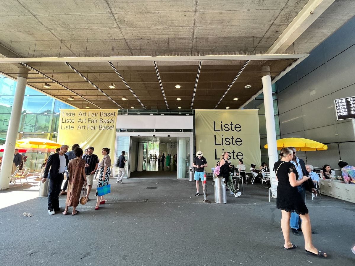 A group of people outside the entryway to a fair with signs reading 'LISTE' outside it.