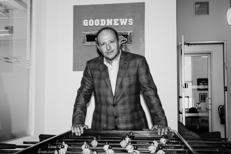 Matt Hiltzik stands behind a foosball table in his office, leaning forward with hands on the table's edge, wearing a plaid blazer. A sign reading "GOODNEWS" hangs on the wall behind him.