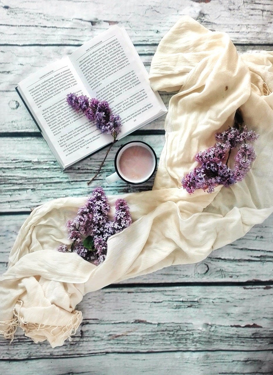 On a white-painted wooden background sits a book that is surrounded by sprigs of purple lilac and a white scarf. Also there is a coffee cup full of a warm drink.