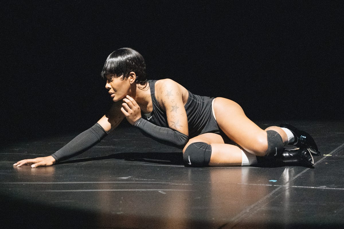 Ms. Boogie, a performer, is captured mid-performance lying on the stage floor. She wears a black sleeveless bodysuit, black knee pads, black gloves, and shiny black boots. Her short, sleek hairstyle and tattoos are highlighted under the dramatic stage lighting. Her pose, with one hand reaching forward and her face turned to the side, conveys intensity and focus against the dark background.