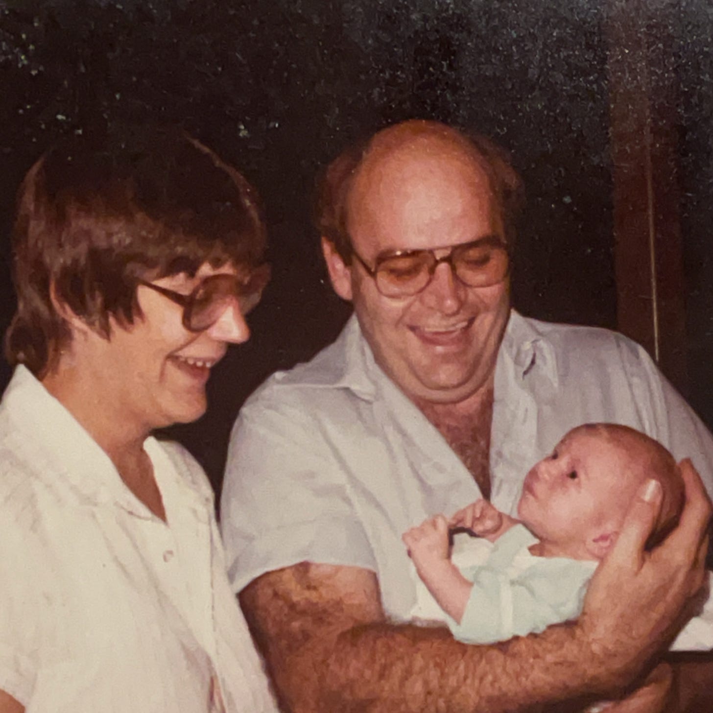 An old photo shows two parents looking delighted at their tiny baby, who is maybe 3 months old.