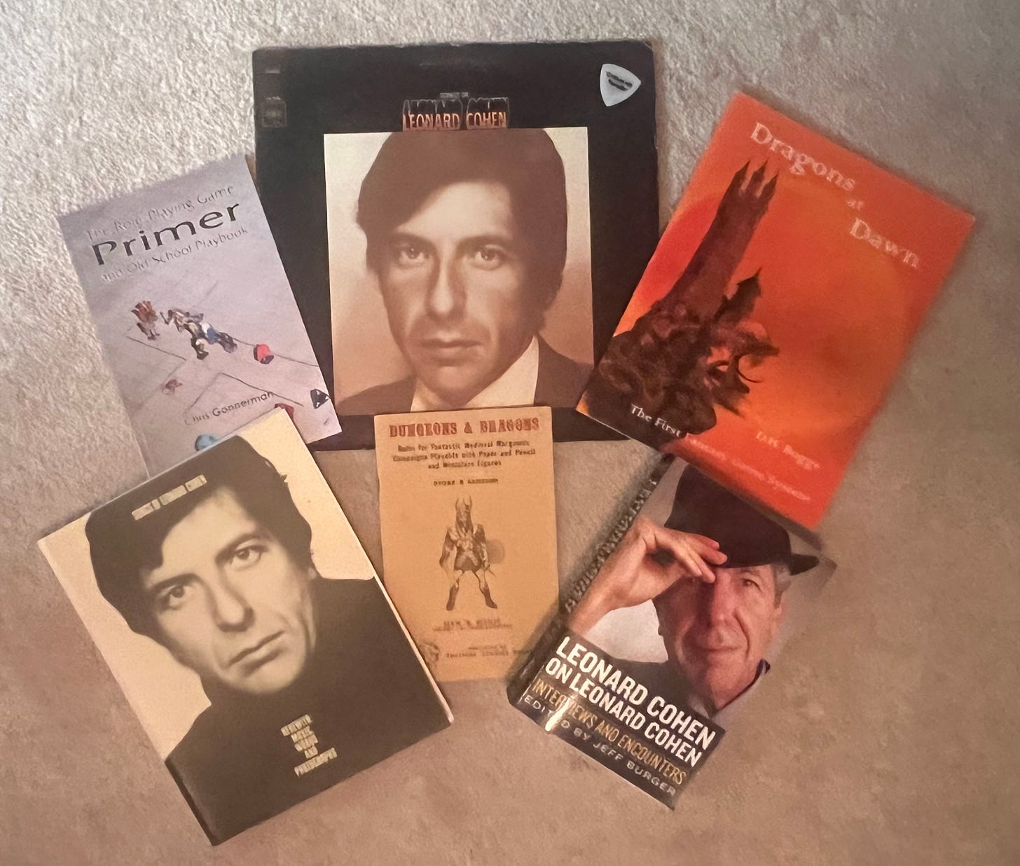A photo of several old school roleplaying game books, two Leonard Cohen books, and a Leonard Cohen album. A white Critical Hit Parader guitar pick lays handsomely on top of a Songs of Leonard Cohen vinyl album, depending on your politics.