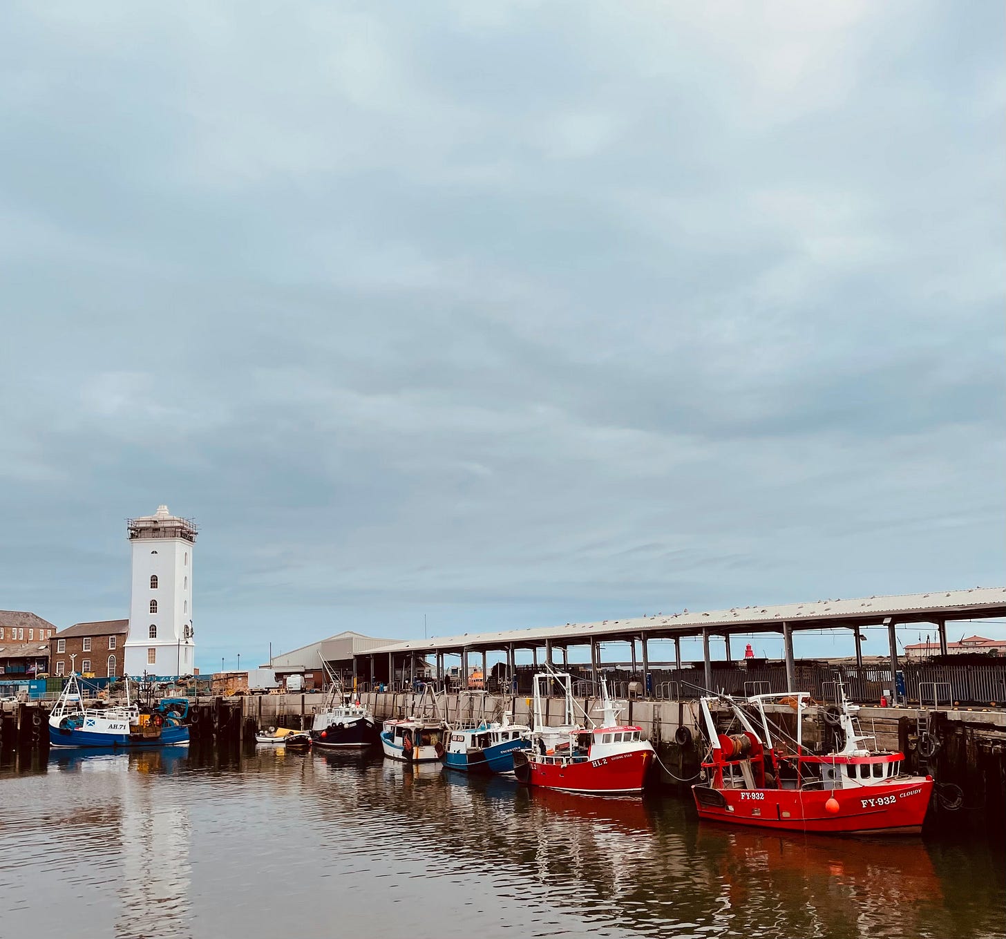 Lots of blueish sky, and in the foreground a harbour where the fishing boats are tied up safely, and a squat, square lighthouse.