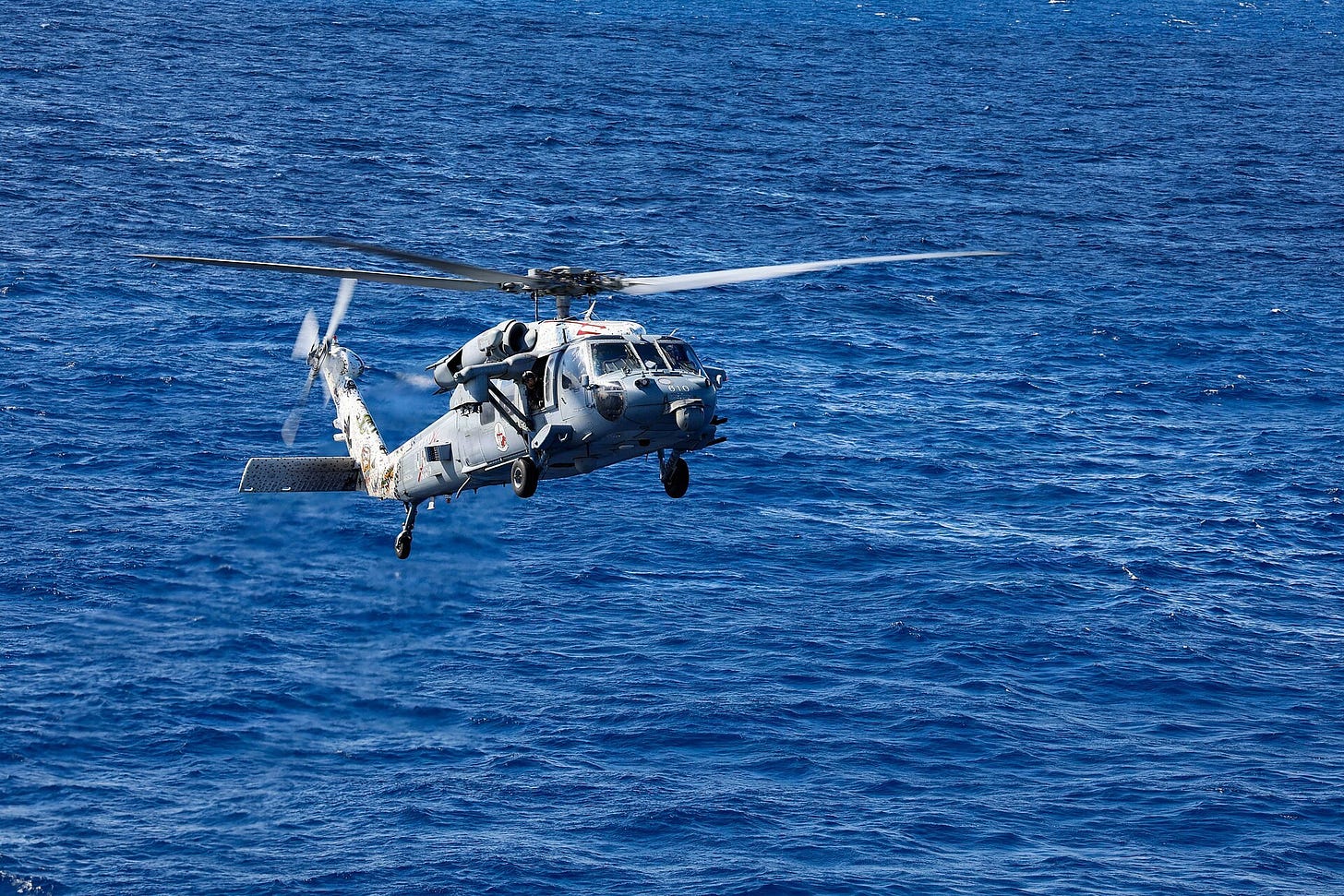 A grey U.S. Navy MH-60S Sea Hawk helicopter flies low over deep blue water as it approaches an aircraft carrier's flight deck. A grey U.S. Navy MH-60S Sea Hawk helicopter flies low over deep blue water as it approaches an aircraft carrier's flight deck.
