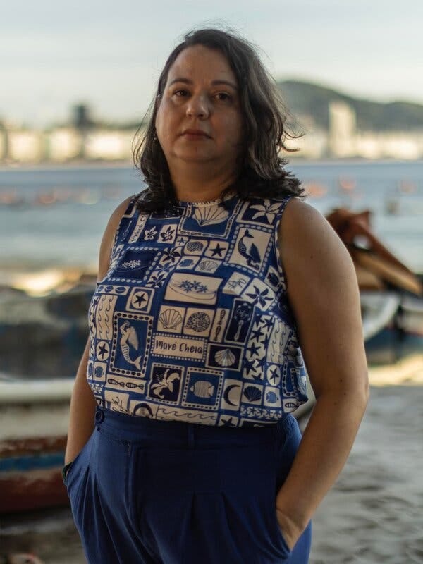 Ana Vaz, in a blue and white blouse decorated with fish and sea shells, stands near a coastline in Rio de Janeiro. 