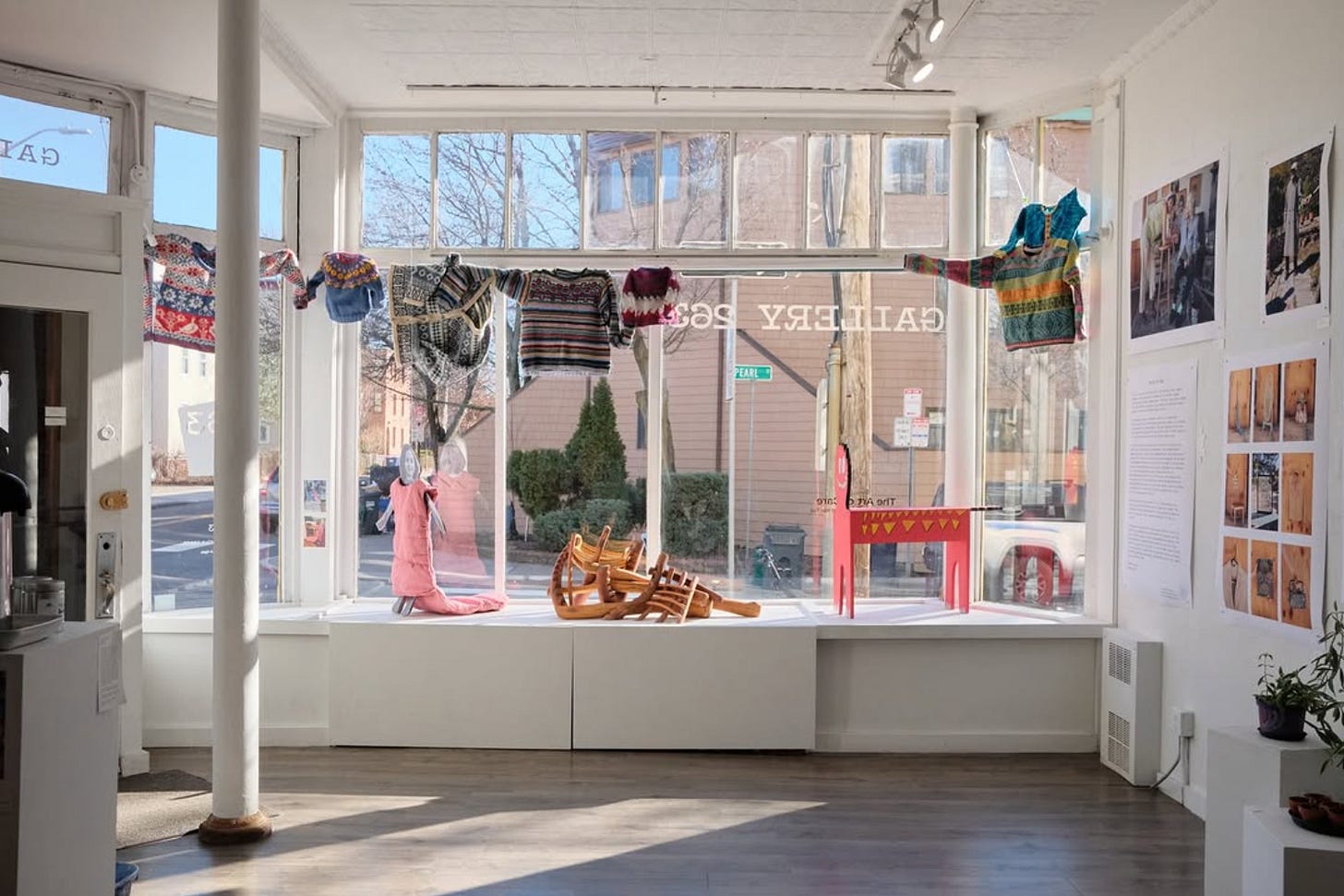 Looking out towards the windows of a gallery, from the inside space. There are sweaters hanging from a curtain rod and photos on the wall.
