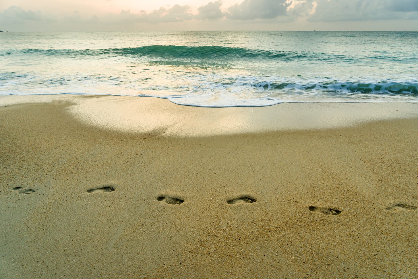 A trail of bare footprints crosses wet golden sand toward a gently breaking turquoise wave, under a soft pink-and-gray sunset sky. A trail of bare footprints crosses wet golden sand toward a gently breaking turquoise wave, under a soft pink-and-gray sunset sky.