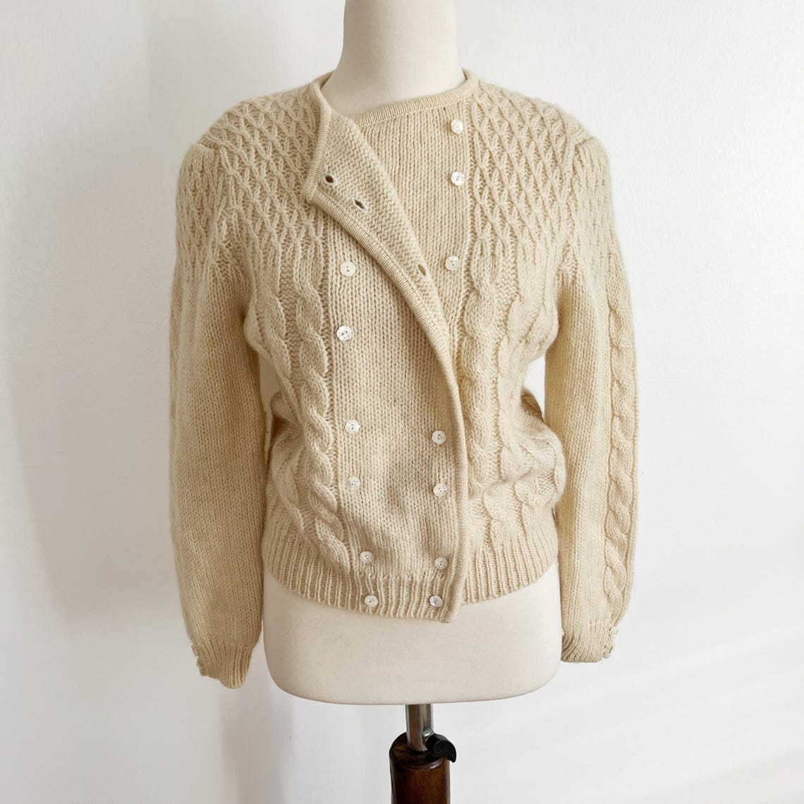 May include: A cream-colored, cable-knit cardigan sweater and matching top. The cardigan has a double-breasted style with faux buttons.  The long sleeves and close-fitting design suggest a vintage or retro style. The sweater set is displayed on a mannequin against a white background.