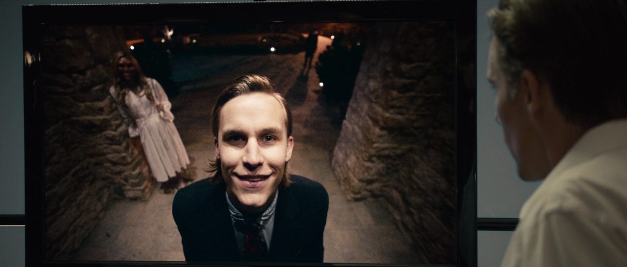 A man watches a creen with security camera footage, in which a well-dressed man with slicked back hair smiles into the camera. A masked woman in a white dress stands in the background.