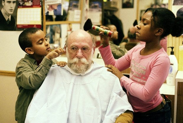 In a barber shop, an older man sits draped in cloth while two school-aged children cut his hair. In a barber shop, an older man sits draped in cloth while two school-aged children cut his hair.