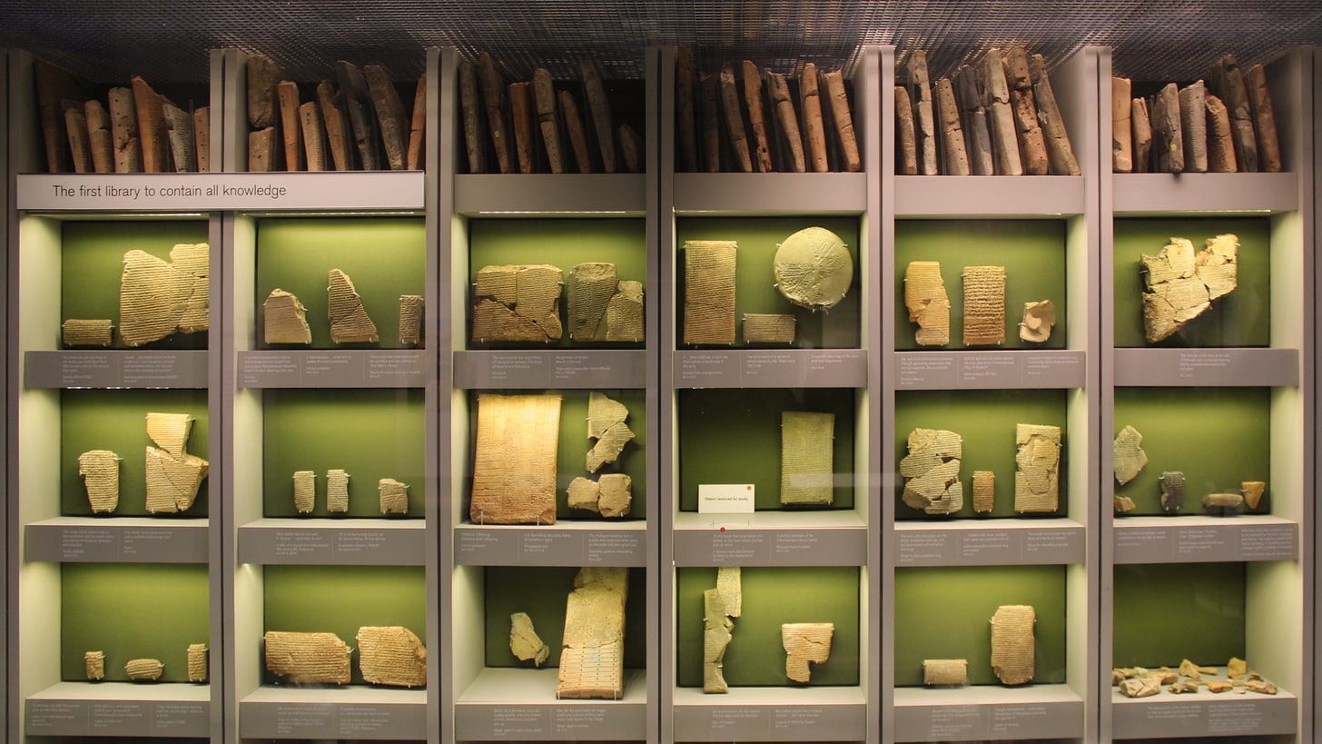 Display case with ancient clay tablets and scrolls, labeled as "The first library to contain all knowledge," arranged on shelves in a museum exhibit.