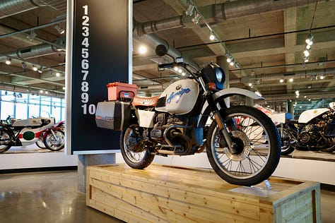 Barber Vintage Motorsports Museum Sign; vintage motorcycles on display; Jessica with her hair in two braids in one of the areas of the bike museum;dozens of bikes stacked on a very tall display; King Kenny Roberts wearing a cowboy hat and sitting on a yellow bike