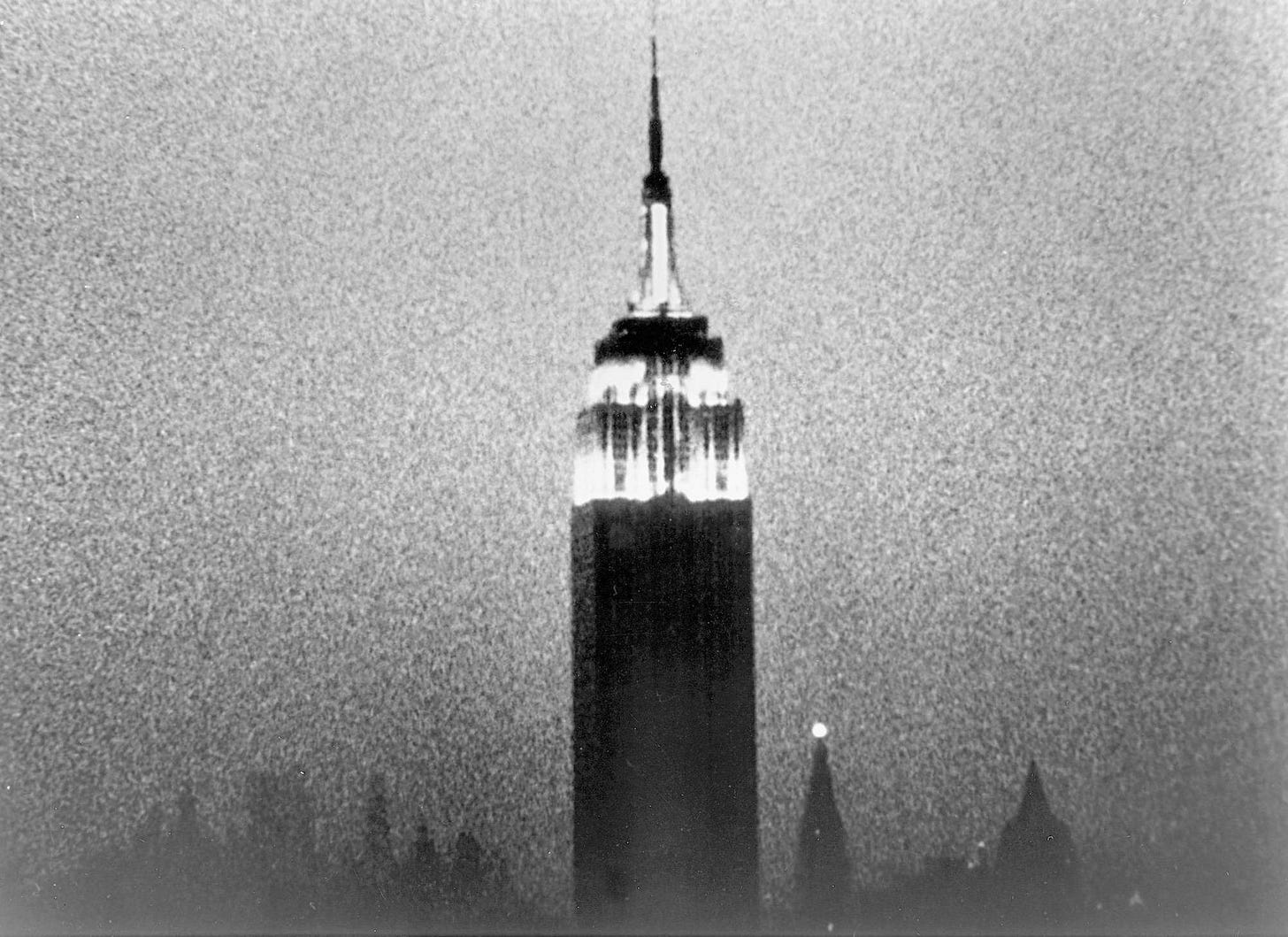 Black and white film still from Andy Warhol and John Palmer's Empire, 1964, showing the Empire State Building illuminated at dusk, against a backdrop of sky clouded with film-grain. At the bottom of the image is the silhouette of the New York skyline.