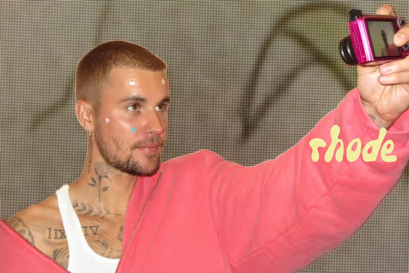 Justin Bieber taking a selfie with a vintage pink digital camera while wearing colorful Rhode Spotwear blemish patches designed for Coachella 2026. Justin Bieber taking a selfie with a vintage pink digital camera while wearing colorful Rhode Spotwear blemish patches designed for Coachella 2026.