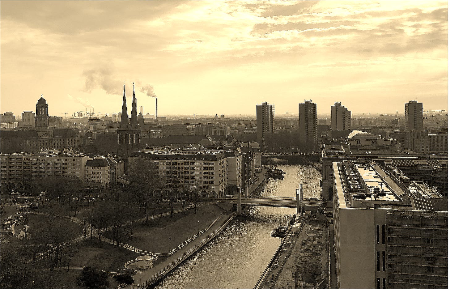 A view over Berlin with church spires, smokestacks, high-rise buildings, and the Spree River under a cloudy winter sky.