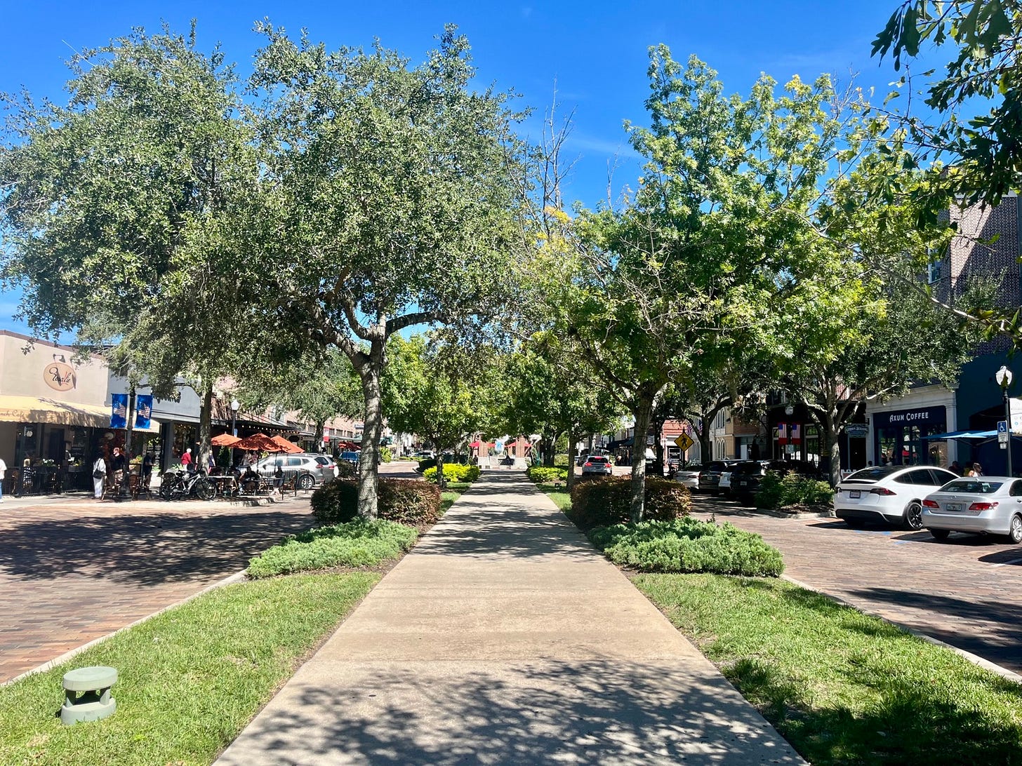 The West Orange Trail runs through downtown Winter Garden, Florida, beneath mature live oaks and between brick-paved streets lined with shops, outdoor dining, and pedestrians.