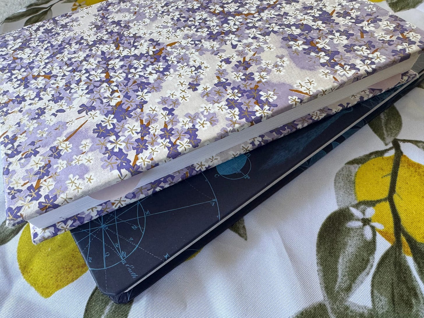 Two journals - top one has purple flowers with gold accents and bottom one is dark blue with mathematical diagrams - sitting flat on a wrinkled tablecloth with lemons
