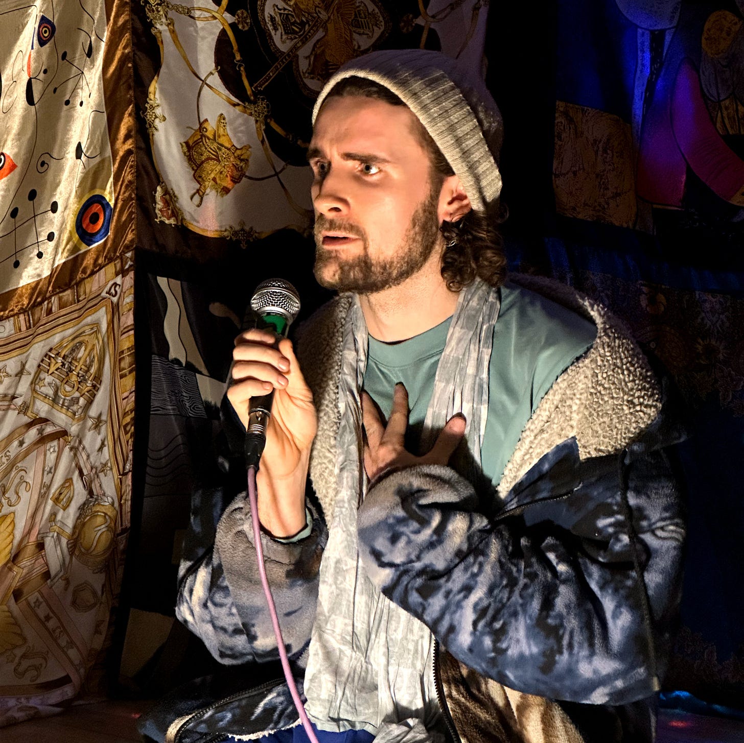 Shaun Hill performing poetry in low dramatic light, holding a microphone with a pink cable, one hand pressed to his chest. He wears a grey beanie, a plush wolf hoody falling off one shoulder, and a scarf. He's surrounded by richly patterned fabric hangings. His expression is intense and mid-speech, eyes looking slightly off-camera.