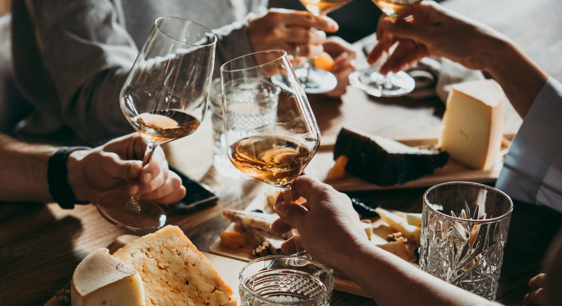 A group of people around a wooden table with cheese on top clinking wine glasses A group of people around a wooden table with cheese on top clinking wine glasses