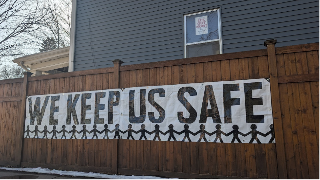 A sign on a brown wooden fence that says We Keep Us Safe and underneath the words outlines of people in a row with hands connected.