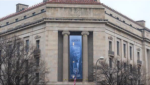 A large, vertical blue banner hangs between neoclassical columns on exterior of the Department of Justice headquarters. A large, vertical blue banner hangs between neoclassical columns on exterior of the Department of Justice headquarters.