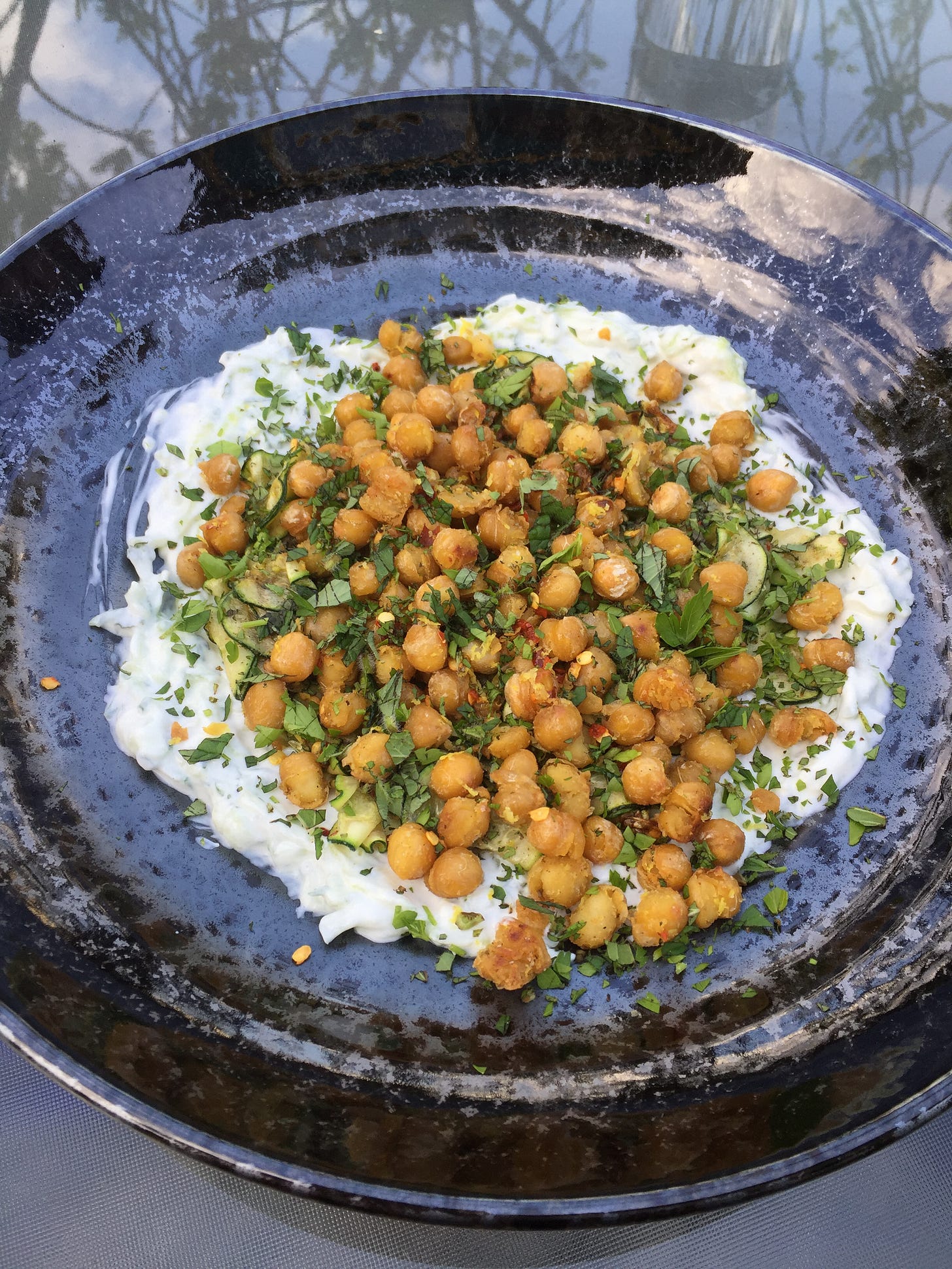 On a glass patio table, a shallow black speckled dish with tzatziki spread in the centre. On top of the dip is sautéed slice of zucchini covered with crispy chickpeas, dusted with herbs and chili flakes.