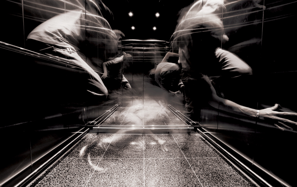 Black and white photo inside elevator with blurred, distorted figures depicting claustrophbia