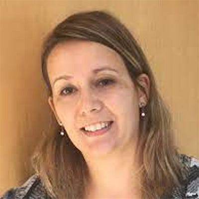 Mulher adulta, de cabelos castanhos e lisos, na altura dos ombros. Ela está sorrindo e olhando para a câmera. Usa brincos pequenos, uma blusa preta e um casaco estampado em tons de cinza e preto. Ao fundo, há uma parede de madeira em tom marrom-claro. Mulher adulta, de cabelos castanhos e lisos, na altura dos ombros. Ela está sorrindo e olhando para a câmera. Usa brincos pequenos, uma blusa preta e um casaco estampado em tons de cinza e preto. Ao fundo, há uma parede de madeira em tom marrom-claro.