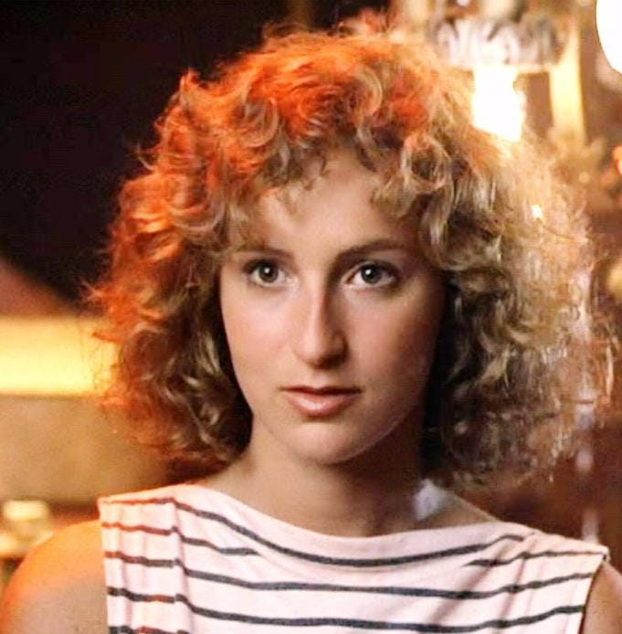 A close-up cinematic film still from the 1980s classic Dirty Dancing movie showing a young Jennifer Grey with natural, fresh-faced makeup and a radiant pink blush flush on her cheeks.