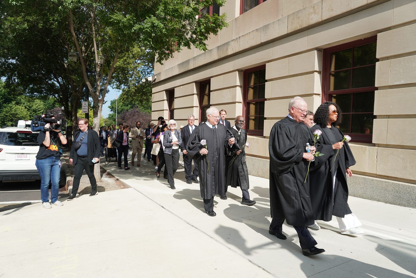 a procession of judges and other people down a sidewalk by the side of a concrete municipal building, wearing robes and each holding a rose a procession of judges and other people down a sidewalk by the side of a concrete municipal building, wearing robes and each holding a rose