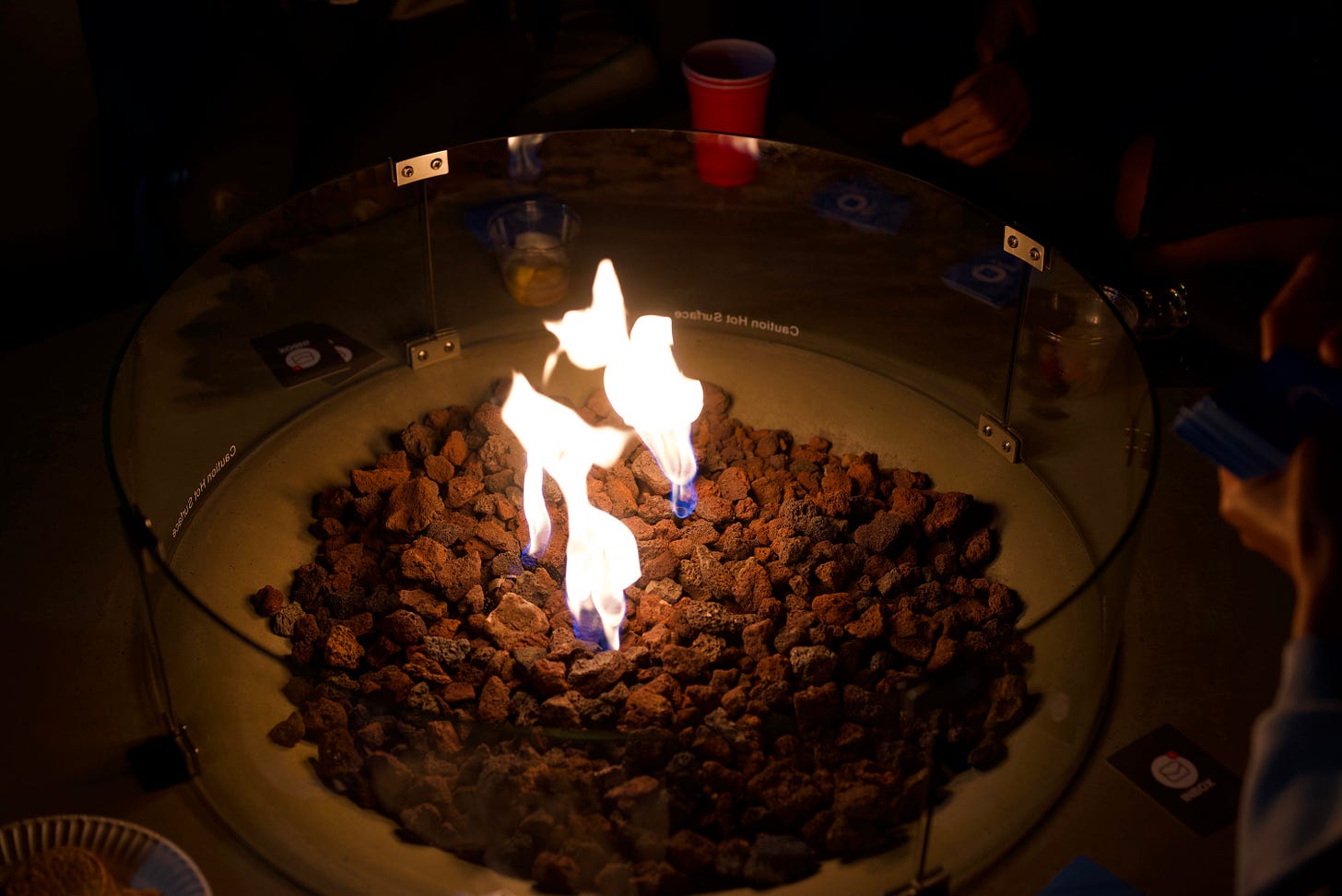 Outdoor fire pit surrounded by glass with flames rising from dark stones while people sit nearby in a dimly lit setting. Outdoor fire pit surrounded by glass with flames rising from dark stones while people sit nearby in a dimly lit setting.