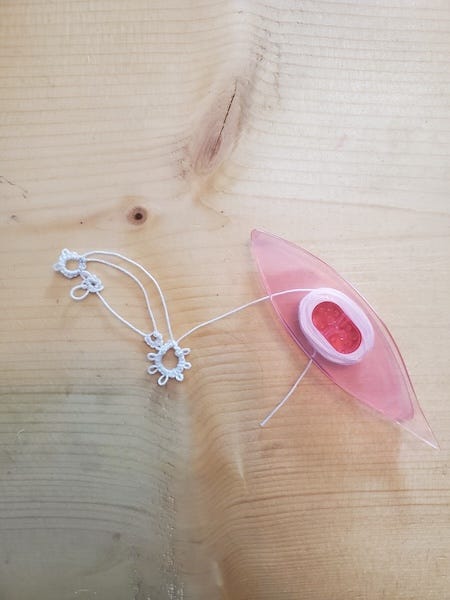 a pink plastic tool--a shuttle--used to make tatted lace displayed on a wood table with a few bits of tatted material